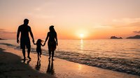 Family with two adults and one child in wetsuits walking into ocean at sunset, holding hands. Golden hour beach scene for travel, lifestyle, and family content.