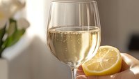 Glass of sparkling white wine with fresh cut lemon slice in soft natural lighting, bubbles rising in golden liquid against blurred bright background.