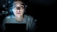 Young Caucasian male programmer working late at night with holographic data interface projections illuminating face in dark room for technology concepts.