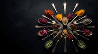 Colorful spices and herbs arranged in wooden spoons forming circular pattern on dark slate background for culinary cooking and seasoning concepts.