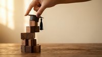 Hand placing graduation cap on wooden blocks tower symbolizing educational achievement and academic success journey on warm background.