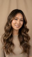 Young Asian woman with long wavy brown hair and highlights smiling warmly in beige sweater against neutral background for beauty and lifestyle content.
