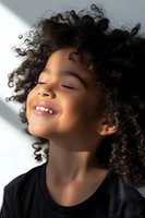 Young African American girl with curly natural hair laughing joyfully with eyes closed, wearing black top, captured in soft natural lighting against light background.