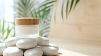 Cream jar with wooden lid on stacked stones, tropical plants background, skincare product display for cosmetics, beauty, wellness branding.