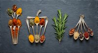 Colorful spices and herbs arranged on wooden spoons with fresh rosemary on dark slate background for culinary cooking and seasoning recipes.