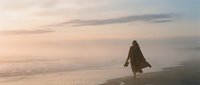 Woman in long coat walking on misty beach at dawn, solitary figure in fog, contemplative mood, suitable for editorial, wellness, and lifestyle design projects.