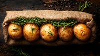 Fresh organic potatoes with rosemary sprigs arranged on rustic burlap sack against dark wooden background, raw farm vegetables for cooking.