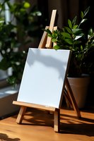 Wooden easel with blank white canvas on wooden table near green plant in sunlight, creating artistic workspace atmosphere with natural shadows.