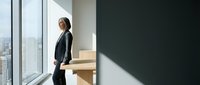 Businesswoman in formal attire standing by large office window with city skyline view, contemplating career decisions and professional growth opportunities.