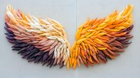 Vibrant angel wings art installation made from layered paper or cardboard pieces in gradient colors from dark purple to white and bright orange, mounted on light wall.