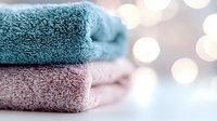 Soft folded bath towels in pastel blue and pink colors on white surface with blurred bokeh lights background, creating cozy spa atmosphere.