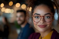 Young African American woman with curly hair and stylish glasses smiling warmly at camera in soft evening lighting, with blurred people in background. Perfect for diversity campaigns.
