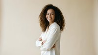 Confident young African American businesswoman with curly hair smiling in white blazer against neutral background for corporate presentations.