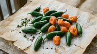 Fresh green and orange hot chili peppers scattered on rustic parchment paper with black pepper seasoning on wooden table for cooking and spice preparation.