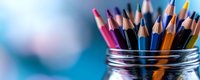Colorful wooden pencils standing in glass jar against blurred blue background, creative art supplies arranged for drawing and sketching activities.