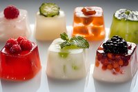 Colorful fruit jelly desserts with fresh berries, cucumber slices, and mint garnish arranged on white surface.