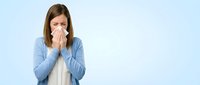 Sick woman with tissue blowing nose against blue background, depicting cold, flu, allergy symptoms, illness, healthcare and wellness concepts.