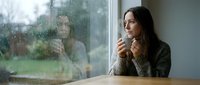 Woman with long dark hair and fair complexion sitting at table gazing through rainy window, holding warm beverage, contemplative mood, suitable for lifestyle, wellness, mental health, seasonal content.