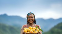 Young woman of African descent wearing a blue headwrap holds a basket of fresh yellow and orange mangoes against a misty mountain landscape backdrop.