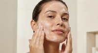 Young Caucasian woman applying white facial cleanser or moisturizer to her face in bathroom, skincare routine for healthy glowing skin.