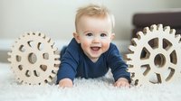 Happy Caucasian baby boy in navy blue outfit lying on white carpet between two large decorative wooden gears, expressing joy and curiosity during playtime.