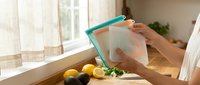 Woman using reusable silicone food storage bag in modern kitchen with fresh vegetables on wooden cutting board for eco-friendly meal prep and organization.