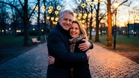 Happy mature Caucasian couple embracing outdoors at sunset with warm bokeh lights creating romantic atmosphere in urban park setting during golden hour.