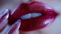 Sensual close-up of glossy dark red lips with metallic shimmer effect and manicured finger touching corner of mouth, dramatic beauty makeup detail.