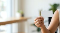 Woman holding blank white business card in modern bright office space with plants and natural light for corporate branding and professional networking purposes.