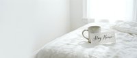 Cozy white bedroom with a ceramic mug and Stay Home card on soft linen bedding, evoking comfort, relaxation, and a peaceful morning at home.