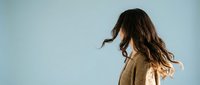 Young woman with flowing brown hair in beige coat against clear blue sky background, hair movement in wind, freedom and lifestyle concept.