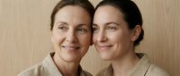 Two Caucasian women of different ages smile warmly together against a wooden background, wearing matching beige tops, showcasing family bond and generational beauty.