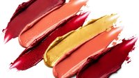 Colorful lipstick smears in warm autumn tones including burgundy coral gold and red creating artistic makeup swatches on white background for beauty cosmetics.