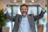Successful mature businessman with raised arms celebrating victory in modern office environment, expressing joy and achievement in corporate setting.
