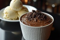 Rich chocolate mousse topped with chocolate shavings in white textured cup, with vanilla ice cream scoops on plate in background.