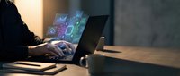 Professional working on laptop with holographic digital interface displaying data analytics and futuristic technology elements in modern office environment.