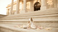 Elegant bridal heels resting on marble steps with scattered rose petals, classical architecture in warm golden light backdrop.