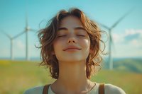 Young woman with closed eyes and serene smile enjoying sunlight at wind turbine farm, curly brown hair moving in breeze against turquoise sky.