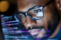 African American male programmer in glasses looking at computer screen with reflected programming code, focused on software development work in dark environment.