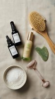 Skincare and wellness products arranged flat lay with natural bristle brush, serums, facial roller, jade stone, and powder on neutral linen background for beauty routines.