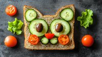 Creative cat-shaped sandwich with avocado, cucumber, olives, and cherry tomatoes on whole grain bread. Fresh vegetables arranged as playful food art.