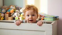 Adorable redhead toddler boy peeking over white toy chest edge in bright nursery room with colorful toys and books creating playful childhood moment.