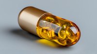 Golden transparent capsule with amber liquid and small white pellets inside, lying on gray background. Pharmaceutical concept for medicine and supplement industry.