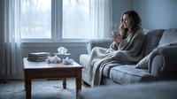 Woman with long dark hair, appearing to be in her 30s, sitting on a gray couch wrapped in a blanket, holding a warm beverage while looking out a window during daytime. Tissues and books on table nearby.