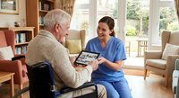 Healthcare worker showing tablet to elderly Caucasian man in wheelchair during home visit consultation in bright living room setting.
