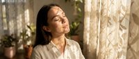 Young Asian woman with closed eyes enjoying warm sunlight streaming through window in peaceful morning meditation moment at home.