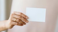 Young woman with medium skin tone holding blank white card against neutral beige background, suitable for business cards, invitations, marketing mockups, and promotional designs.