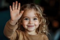Cheerful Caucasian girl with curly blonde hair wearing beige sweater waves hello with bright smile against dark background. Genuine childhood joy and friendliness.