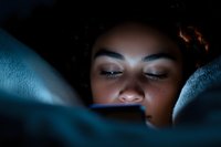 Young African American woman illuminated by phone screen light while lying in bed at night, creating moody atmosphere with blue tones and dramatic shadows.