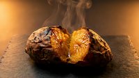 Hot baked potato split open with steam rising, golden flesh exposed on dark wooden surface with warm lighting for culinary and food photography.